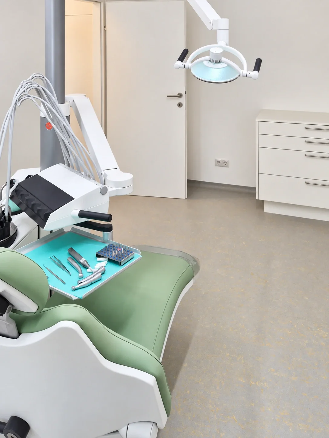 Seitliche Ansicht einer gr&uuml;nen zahn&auml;rztlichen Behandlungseinheit mit gro&szlig;er schwenkbarer Leuchte; daneben ein wei&szlig;er Schubladenschrank und eine offene T&uuml;r.