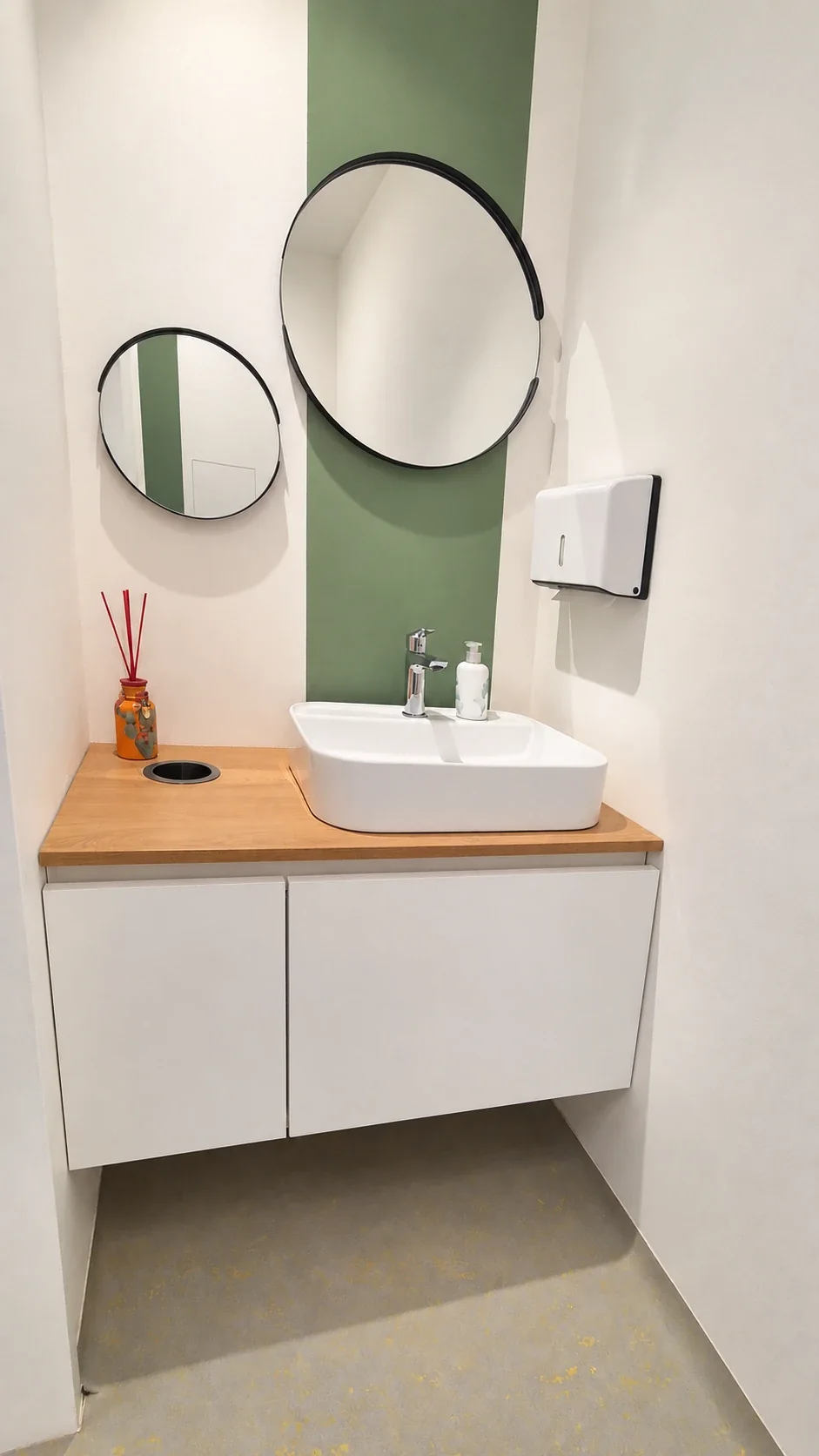 Wei&szlig;es Aufsatzwaschbecken auf einer Platte in Holzoptik &uuml;ber einem wei&szlig;en Unterschrank; zwei runde Spiegel h&auml;ngen vor einem senkrechten gr&uuml;nen Wandstreifen.