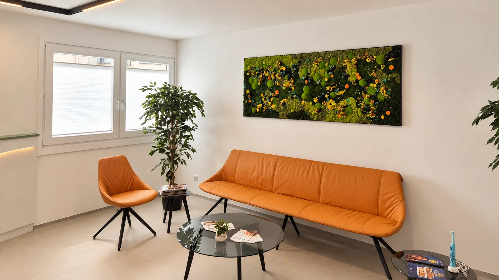 Wartebereich mit ockerfarbener Sitzbank, schwarzen runden Tischen und Pflanzen; ein rechteckiges gr&uuml;nes Pflanzenbild h&auml;ngt an der wei&szlig;en Wand.